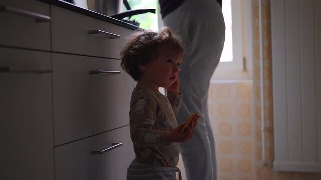 Toddler leaning against mother while she prepares food in family kitchen, highlighting attachment, security, emotional connection, caregiving and everyday family life