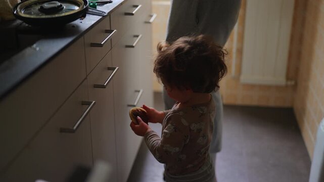 Toddler standing in kitchen holding snack while waiting near countertop at home, highlighting childhood independence, curiosity, everyday family life and self directed behavior