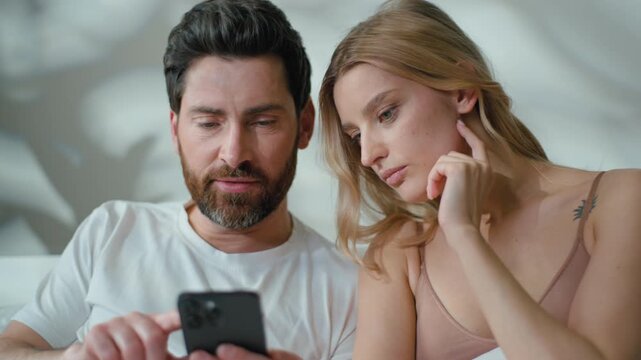 Thoughtful family Caucasian couple thinking hesitate with mobile phone problem talking worried man with woman think browsing smartphone problem error finding solution smiling happy laughing in bed
