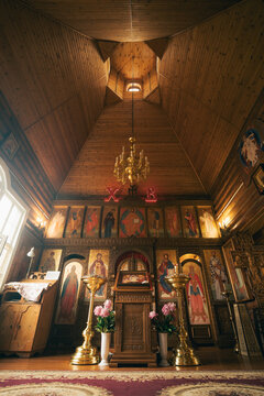 Warm wooden Orthodox church interior with gilded chandelier, elaborate iconostasis, carved altar and candle stands, sunlight from window and skylight lighting icons and sanctuary