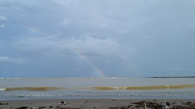 Muddy yellowish ocean waves rolling on the beach with a soft rainbow appearing under a dramatic cloudy sky after a rainstorm