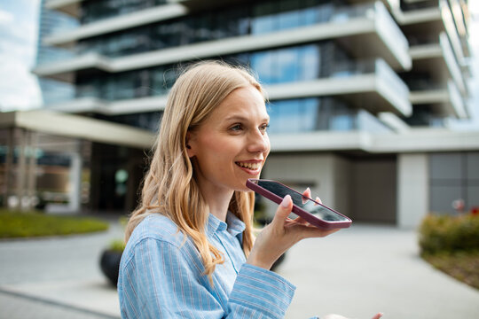 Smiling young woman using voice assistant outside modern apartment building