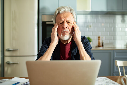 Senior man with headache using a laptop in home kitchen