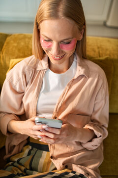 Smiling woman with under eye patches using smartphone at home on sofa