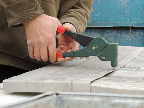 The technician holds a hand-operated lever riveter and squeezes the tool's handles to secure the fastener. The connection is made on curved sheets of thin gray metal.