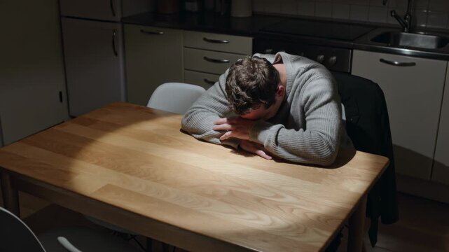 Leaning man in sweater beginning resting head on forearms at kitchen table, jacket on chair