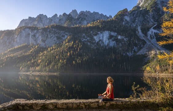 A woman doing yoga by the lake. Lotus position (Padmasana) . Vorderer Gosausee in autumn with a view of the Dachstein mountains. The Gosaukamm on the right. Blue sky, good weather. Reflection. Morning atmosphere. Vorderer Gosausee, Gosau, Gosau valley, Salzkammergut, Upper Austria, Austria