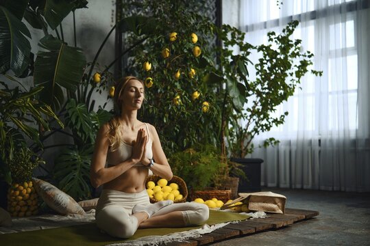 A woman sits cross-legged on a mat with her hands in a prayer position. She is surrounded by plants and lemon fruit. Light comes through the windows, creating a warm atmosphere