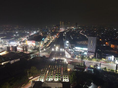 Aerial view of the illuminated highway overpass and urban cityscape at night with traffic streaks and surrounding buildings in Bekasi Barat, West Java, Indonesia.