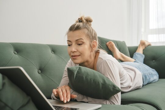 Middle-aged woman lounges comfortably on a green sofa, propped up on a pillow, as she works on her laptop. Sunlight filters through a window, creating a cozy atmosphere in the afternoon
