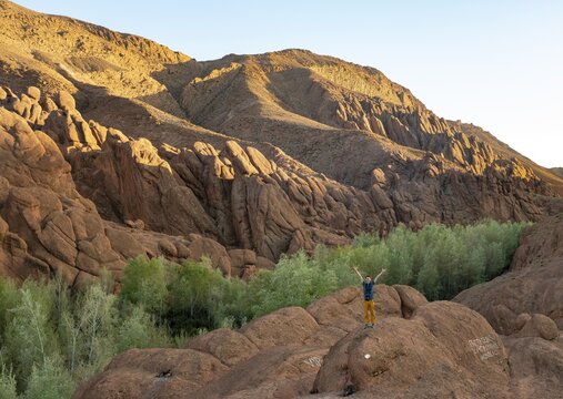 Young man stands on rock and stretches his arms into the air, rock formation Pattes Des Singes, red sandstone rocks, Gorges du Dades, Dades Gorge, Tamellalt, Morocco