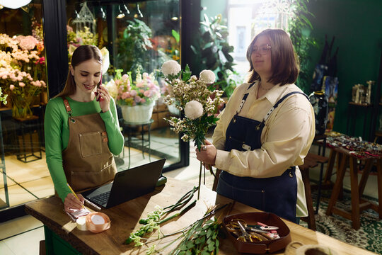 Young adult women managing flower shop orders, arranging bouquet, taking phone call, writing notes beside laptop. Ideal for small business, retail service, local commerce marketing