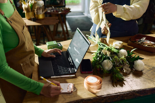 Close up of women florists managing bouquet order, typing on laptop and measuring ribbon at worktable. Useful for small business, ecommerce, retail workflow marketing