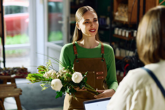 Young adult Caucasian florist holding bouquet and communicating with female customer at checkout in flower shop. Useful for retail service, small business marketing, local commerce promotion