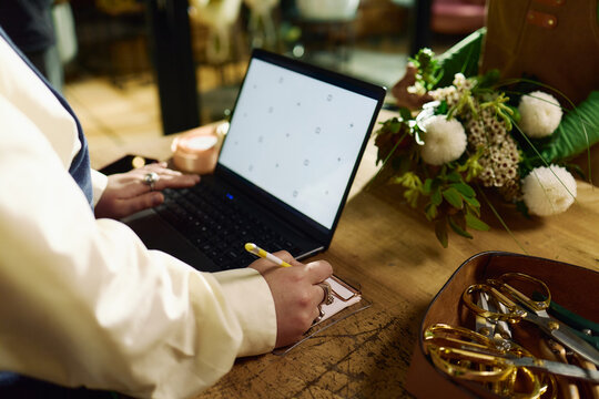 Close up of unrecognizable woman planning floral order, writing notes beside laptop at worktable. Ideal for small business, ecommerce, creative workflow, entrepreneurship marketing