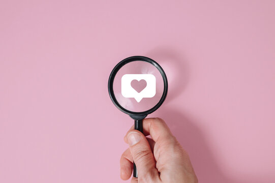 Hand holding magnifying glass over social media heart sign with icons on pink background, digital marketing and engagement research