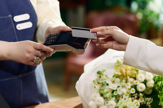 Close up of young adult woman paying with bank card at checkout while florist processing contactless purchase beside bouquet. Ideal for retail payment, small business, ecommerce