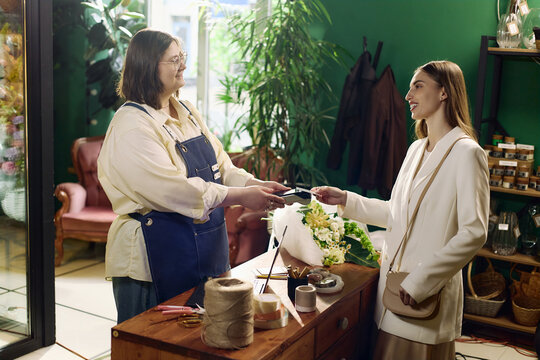 Young adult shop assistant processing card payment from young adult woman at flower store counter, supporting local retail commerce and customer service marketing use
