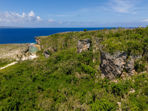 Aerial view of Chigit Beach with rugged limestone cliffs, dense tropical vegetation, and turquoise waters along the coast in Northern Tinian, Tinian, Northern Mariana Islands.