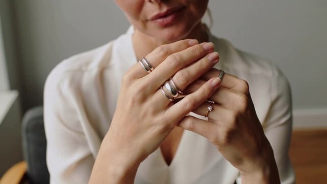 Holding hands, adult woman adjusting and displaying silver rings in quiet room, with white blouse