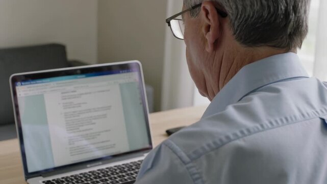 Reading man in blue shirt and glasses, reviewing document appearing on laptop at home with phone