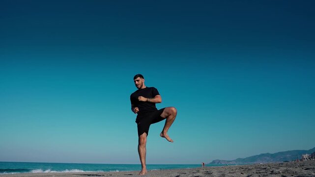 A man performs various martial arts moves barefoot on a sandy beach with mountains and ocean in the background under a clear blue sky.
