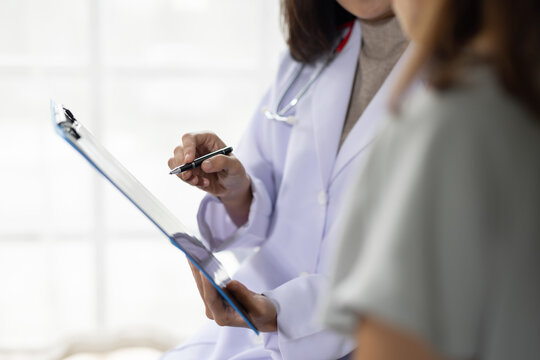 Doctor consulting an Asian woman patient while explaining medical test results or diagnostic information on a clipboard in a hospital or clinic.