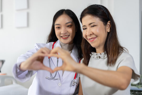 Asian doctor and senior patient expressing love and care, creating a heart gesture with hands, symbolizing a strong bond, empathy, and health support in a clinic.