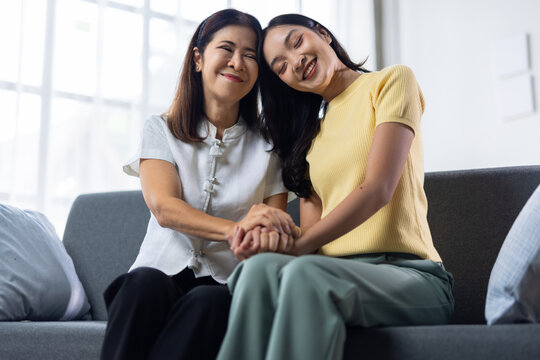 Asian senior mother and adult daughter sitting together on a grey sofa at home, hand in hand, sharing a precious moment of love, support, connection and emotional bond.