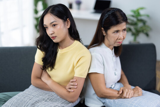 Upset young Asian woman and her mother sit back-to-back on a sofa with arms crossed, illustrating intergenerational conflict, silent disagreement and strained family communication.