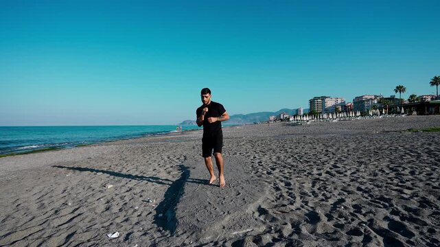 A barefoot man wearing black sportswear practices boxing punches and footwork on a sandy beach with ocean and buildings in the background.
