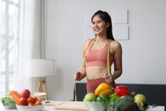 Young woman in sportswear smiling, holding a measuring tape and looking away, surrounded by fresh raw vegetables and fruits, representing healthy eating, fitness lifestyle and weight loss.