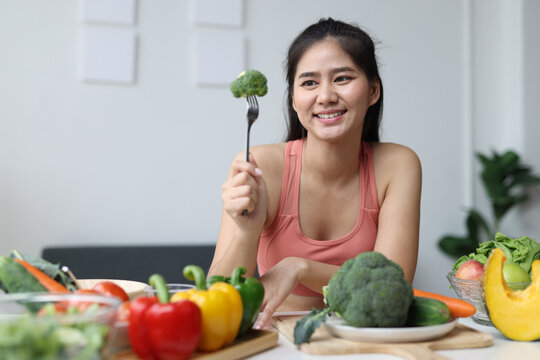 Young Asian woman smiling, holding a fork with broccoli, enjoying a healthy diet with various organic fresh vegetables on a table, wellness and balanced lifestyle.
