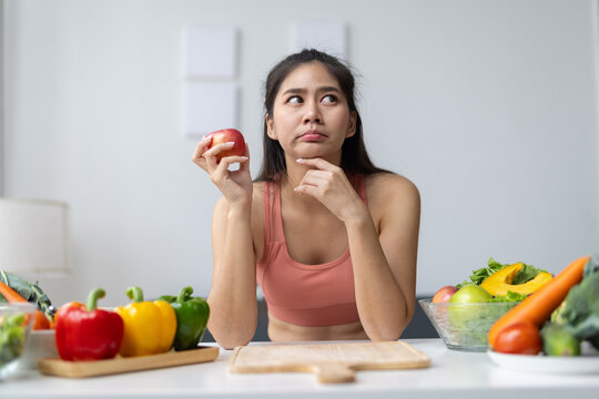 Young Asian woman holding an apple, contemplating her food choices for a healthy diet, surrounded by fresh vegetables on a kitchen counter.