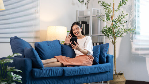Young Asian woman smiling, waving hand while making a video call or taking a selfie on her mobile phone at home.