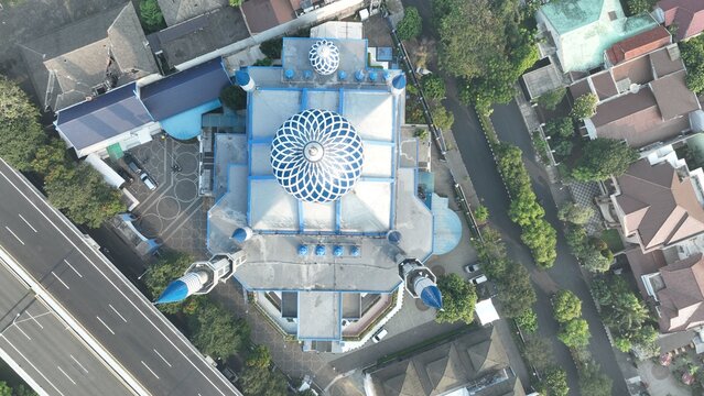Aerial view of a mosque with a distinctive blue and white dome and minarets surrounded by urban rooftops and a nearby highway in Bekasi Barat, West Java, Indonesia.
