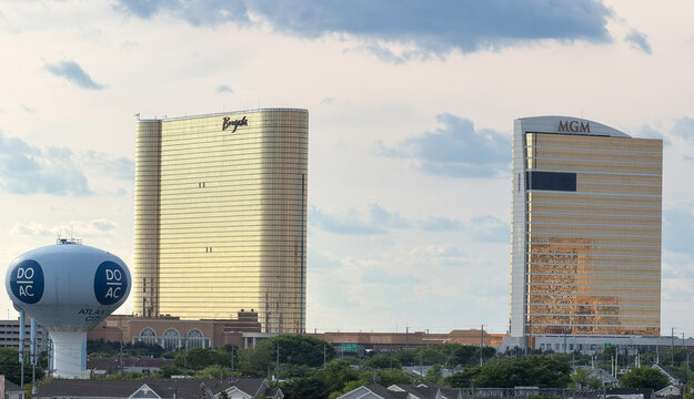 Atlantic City skyline featuring the golden Borgata and MGM casino towers beside a DO AC water tower, reflecting sunlight with clouds above. Urban resort scene conveying tourism, leisure and luxury.