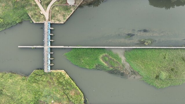 Aerial view of a concrete dam structure spanning a river, with lush green vegetation and water flowing through the sluice gates in Bekasi Barat, West Java, Indonesia.