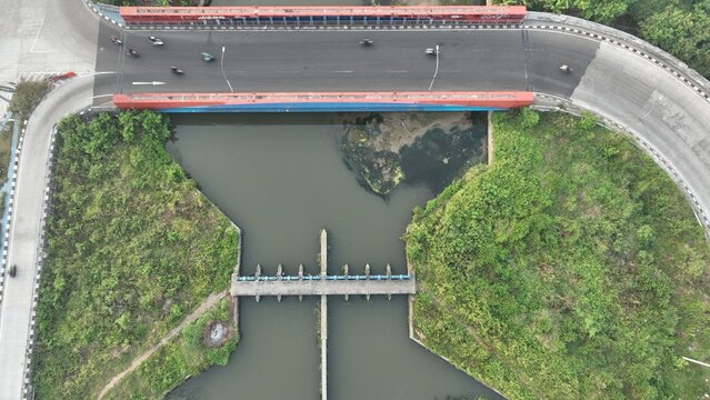 Aerial view of a road bridge crossing a river with a weir structure, surrounded by lush green vegetation in Bekasi Barat, West Java, Indonesia.