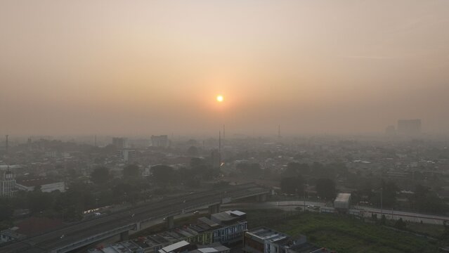 Aerial view of the urban landscape of Bekasi Barat at sunset with a hazy atmosphere, visible highway, and dense rooftops in Bekasi Barat, West Java, Indonesia.