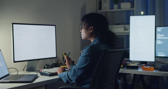 Young African American female programmer coding AI software on multiple screens in night office. Network security, cybersecurity, machine learning, working late, overtime, robotics automation concept.