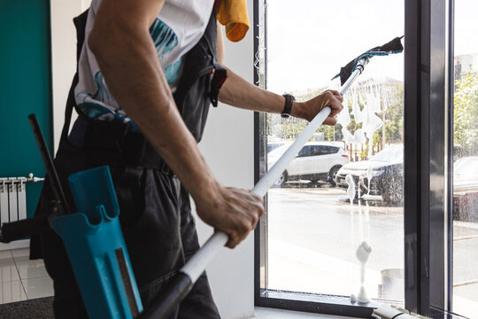 Professional man cleaning large windows with squeegee and pole indoors.