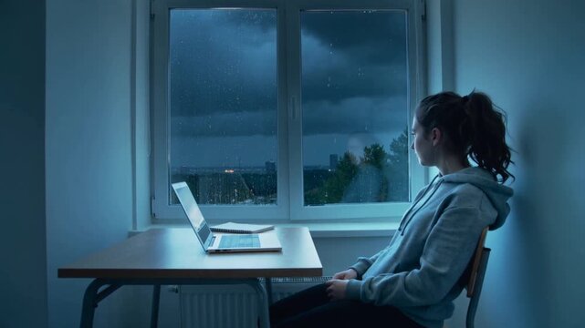 Noticing rain, student pausing laptop at desk and watching storm past window in hoodie, copy space