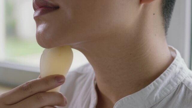 Placing oval tool behind ear, woman using right hand shirt gliding jawline by window, skincare