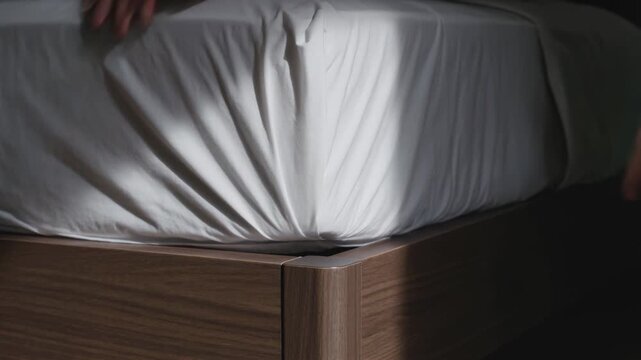 Pulling bare forearms tucking white fitted sheet around mattress corner on wooden bed, making bed