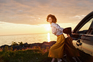 Obraz premium A young woman with curly hair leans on a dark car at sunset by a rocky coastline, conveying a moment of travel and quiet confidence.