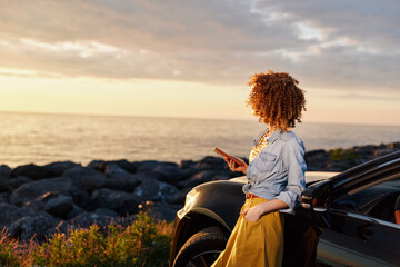 Fototapeta premium Candid woman lifestyle outdoors at sunset by the sea, leaning against a car, denim shirt, yellow skirt, relaxed pose, warm light, gentle breeze, soft colors, horizon glow