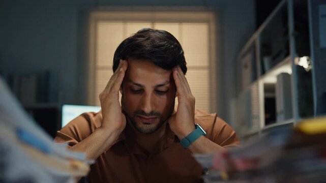 Stressed young Latin businessman having severe headache and migraine at desk in night office. Debt crisis, corporate burnout, late overtime workload, mental health anxiety, business failure concept.