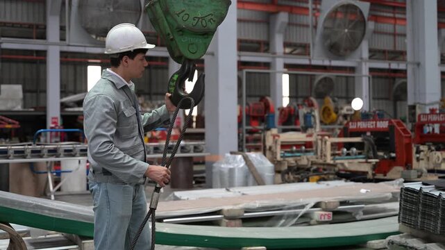An industrial worker wearing protective gear guides and secures a green crane clamp hook mechanism over steel materials in a factory.