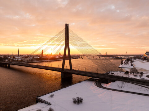 Aerial view of the Vansu Bridge spanning the Daugava River during a golden sunset over the snow-covered city of Riga, Latvia.